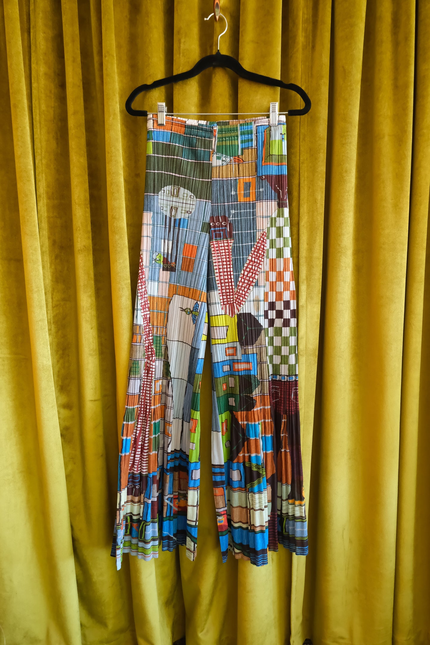 Pleats Please Issey Miyake 2001 archival print flare pants on hanger: front view of both legs showing full multicolor geometric graphic print against gold velvet curtain backdrop, 2001